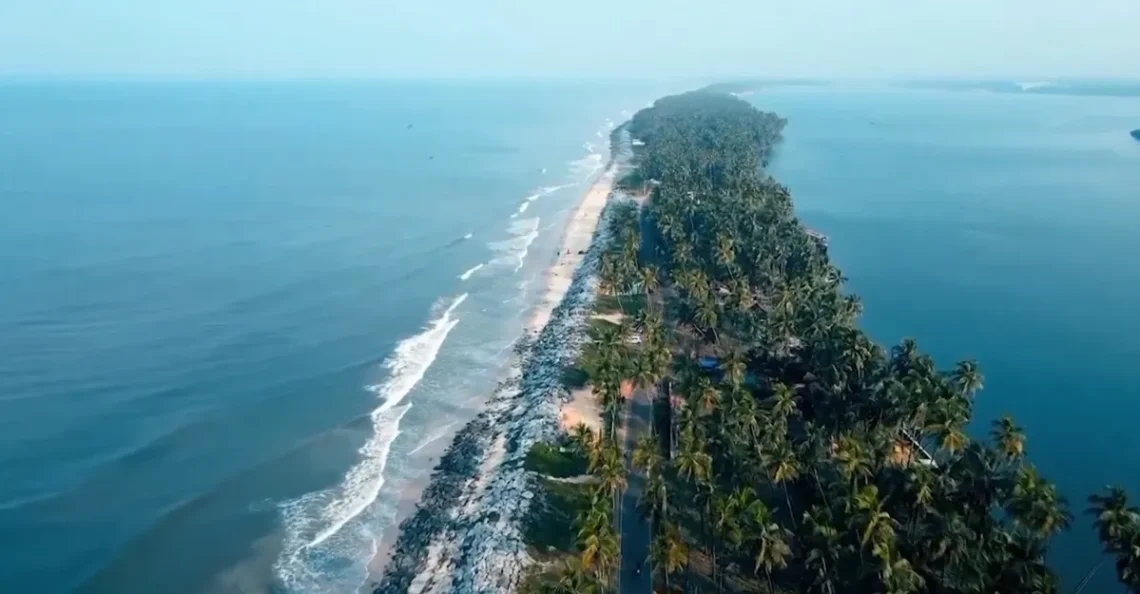 Aerial view of the marvellous Kappil Beach, where a narrow strip of land covered in palm trees stretches between two large bodies of water. Waves crash against the shore on the left, while calmer waters grace the right. A road lined by dense greenery runs along this serene landscape—life is indeed better at the beach.