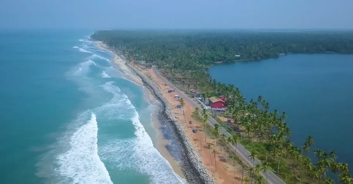 Aerial view of Marvellous Kappil Beach—an idyllic coastal strip flanked by lush coconut trees, with a road running through. Waves crash along the sandy beach on one side, while calm waters are visible on the other. Scattered buildings, including a red-roofed structure, add to its charm.