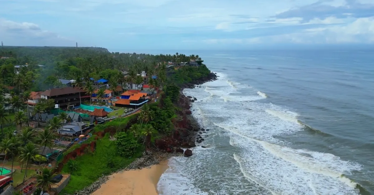 A coastal scene at Varkala Beach, Kerala, shows waves crashing against a rocky shore lined with palm trees and colorful buildings. A sandy beach lies at the edge of the rocks, with greenery and more buildings in the distance under a partly cloudy sky—proof of why life is better at the beach.