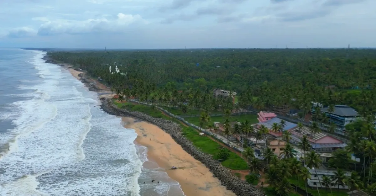 Aerial view of Varkala Beach in Kerala, with waves crashing onto the sandy shore. Dense greenery and palm trees border the coastline, with a few buildings scattered along the edge. The overcast sky adds a dramatic touch. Why is life better at the beach? Just look at this serene landscape!