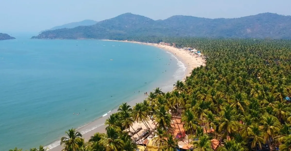 Aerial view of Palolem Beach, Goa, with a crescent-shaped sandy shoreline lined with lush green palm trees. The blue ocean meets the beach with gentle waves, and hills rise in the background under a clear sky. Small buildings dot the coastline near the trees—why life is better at the beach!