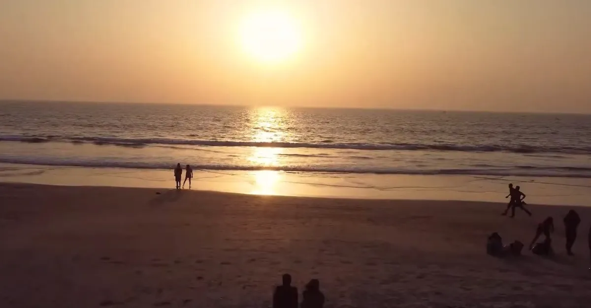 A serene beach scene at Colva Beach at sunset, with the sun setting over the horizon casting a warm golden glow on the water and sand. Silhouetted people are walking and sitting on the beach, enjoying why life is better at the beach in this tranquil scenery.