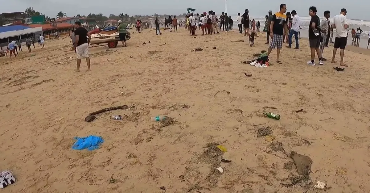 A view of Baga Beach in Goa, featuring a sandy shore with scattered litter, including plastic bottles and bags. Several people are present, some walking and others standing or sitting on the beach. In the background, there are beach shacks and a boat on the sand. The sky is overcast.