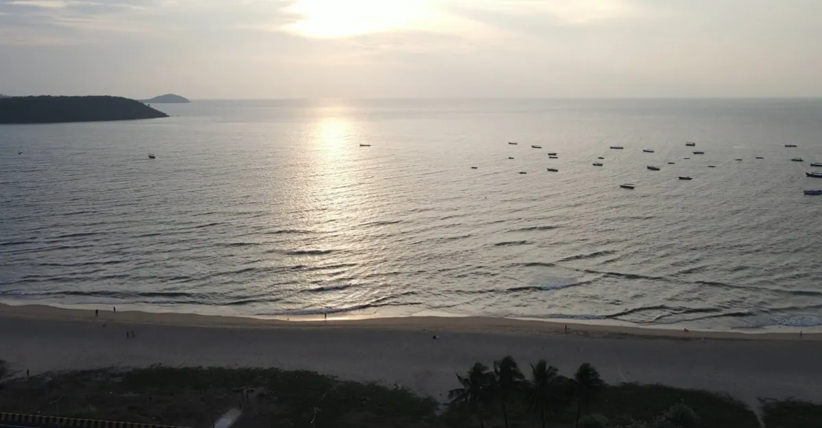 A tranquil view of Baina Beach in Goa during sunrise or sunset. The expansive beach features gentle waves lapping at the shore, with several boats anchored in the calm sea. A landmass is visible on the left side, possibly an island or continuation of the coastline. The sky is mostly clear with subtle light gradients near the horizon, and a few individuals are seen on the beach, appearing small against the vast seascape.