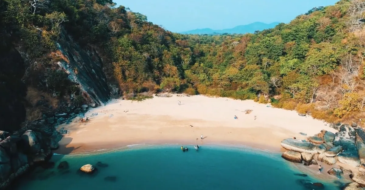 A scenic view of Butterfly Beach in Goa, India. The beach is nestled between lush green hills with dense vegetation. The golden sand is partially shaded by the surrounding hills, creating an inviting secluded cove. A few people and boats are visible on the beach, suggesting it’s a place for relaxation and recreation. The water is calm with gentle waves lapping at the shore, conveying a sense of tranquility and natural beauty.