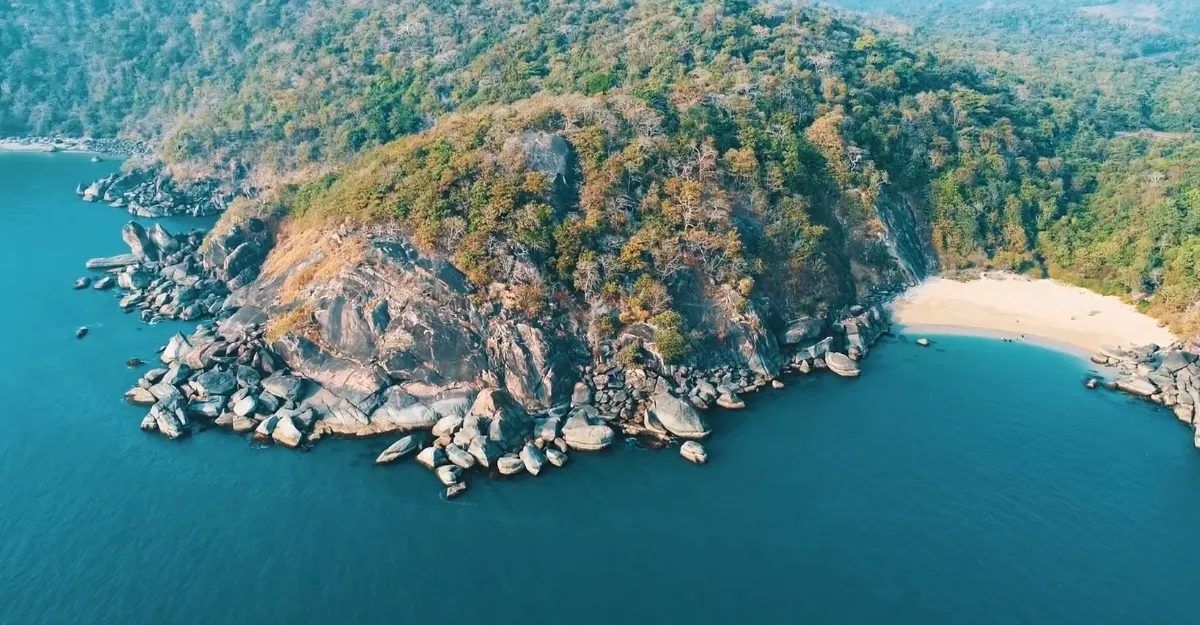 An aerial view of Butterfly Beach in Goa, India. The beach features a semi-circular shoreline with golden sand, flanked by large rocks and boulders along the water’s edge. The calm Arabian Sea gently laps at the shore, and the beach is surrounded by dense forest, adding to its secluded and serene atmosphere.