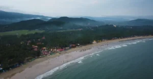 Aerial view of Palolem Beach in Goa, showcasing its sandy shores lined with palm trees and colorful beach shacks. The calm sea with gentle waves meets the shore, while mist-covered hills rise in the background, creating a tranquil and scenic atmosphere.