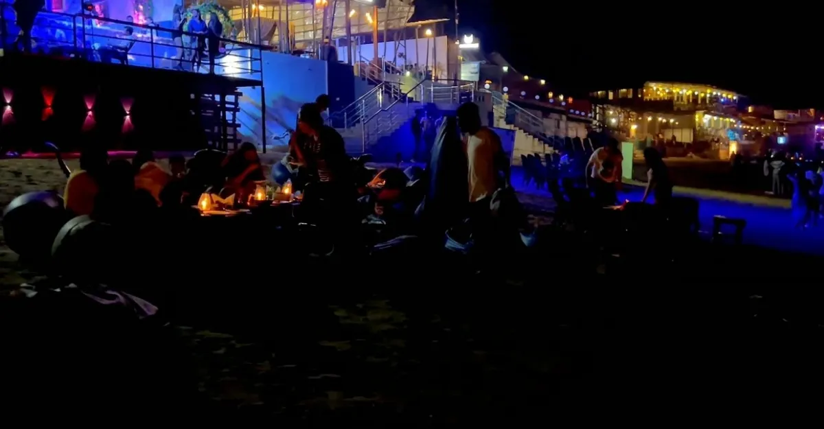 A nighttime scene at Anjuna Beach, Goa. Groups of people are sitting on the sand, some around small fires. The beach is lined with illuminated structures, likely restaurants or clubs, with lights reflecting off the water. The dark sky and warm glow from the fires create a lively and social atmosphere.