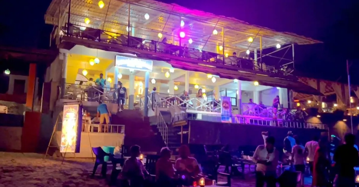 A lively nighttime scene at Anjuna Beach, Goa. The image features a two-story establishment with colorful lights on the upper deck, where people are standing and sitting. The lower level appears to be a restaurant or bar with patrons seated at tables. In the foreground, more people are gathered on the beach, some standing and others seated around candle-lit tables. The dark sky and ambient lighting create a vibrant, social atmosphere by the sea.