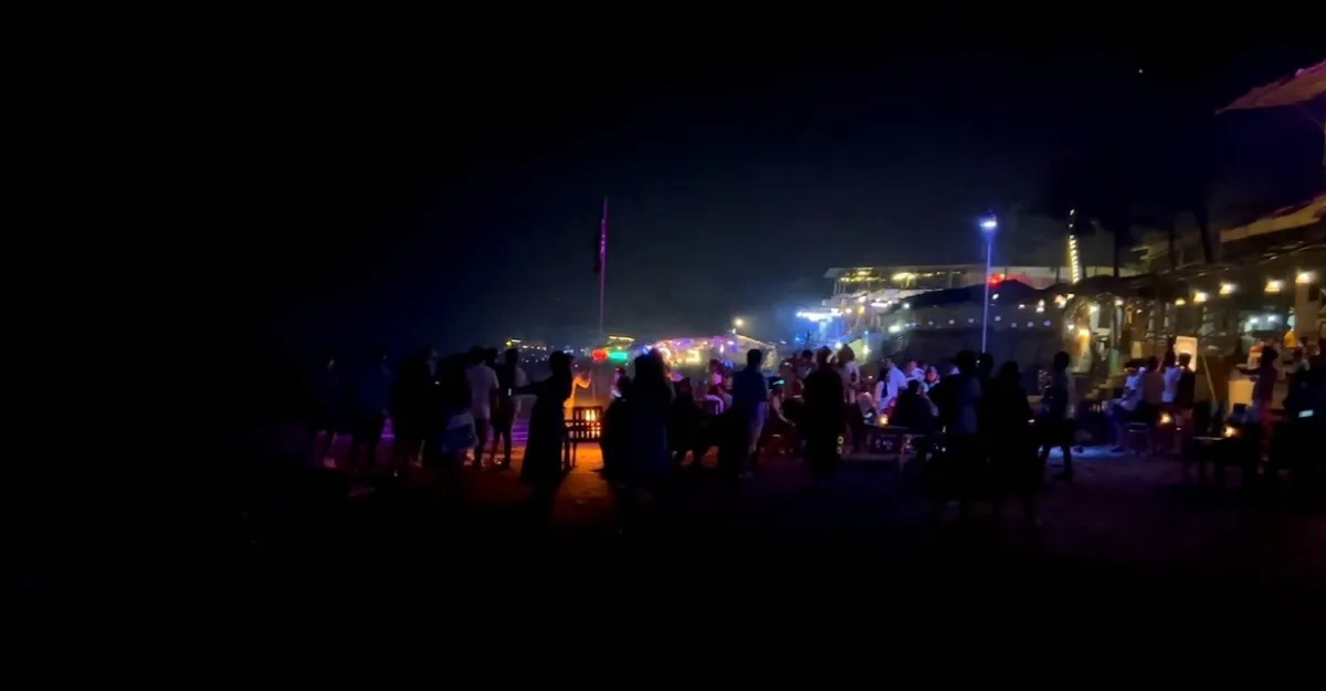 A night scene at Anjuna Beach, Goa. Groups of people are gathered on the sandy beach, some standing and others sitting. The atmosphere is lively with ambient lighting from neon lights and small fires. The background features illuminated buildings, likely beachside establishments. A flag is visible in the center, and the dark night sky contrasts with the vibrant activities below.