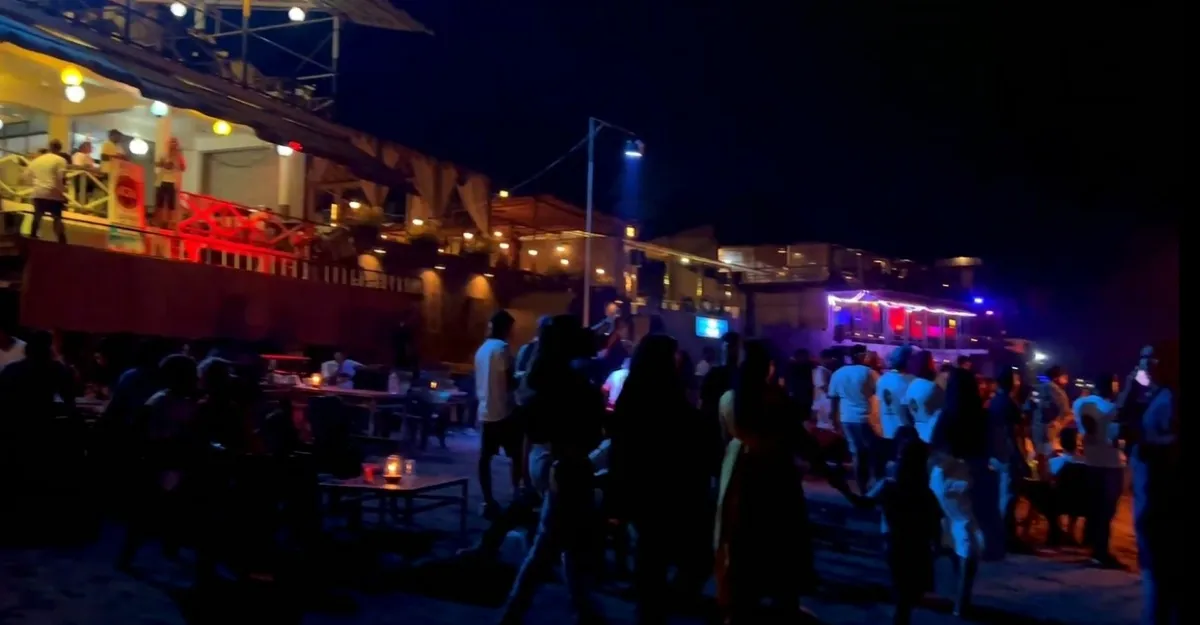 A bustling night scene at Anjuna Beach, Goa. Groups of people are gathered, some standing and others sitting at tables. The background features illuminated buildings, likely restaurants or clubs, with vibrant red and blue lighting. The dark sky indicates it is nighttime, highlighting the lively nightlife atmosphere.