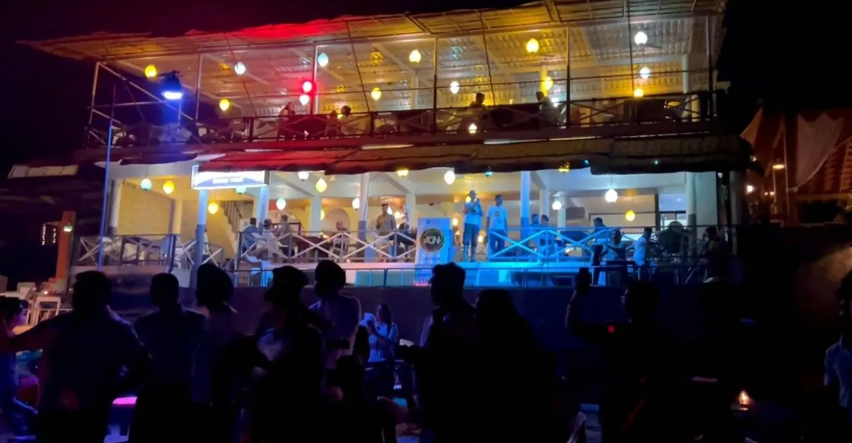 A lively evening scene at Anjuna Beach, Goa. The image shows a two-story open-air structure with bright lights and people on the upper deck. The lower level appears to be a bar or restaurant with patrons standing at the railing. In the foreground, silhouettes of several individuals are seen, suggesting a crowd gathered, possibly watching a performance or enjoying the beach nightlife. The varied lighting in reds, blues, and yellows creates an inviting and vibrant atmosphere.