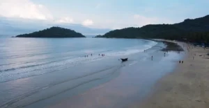 Panoramic view of Palolem Beach in Goa, featuring a gently curving shoreline with light-colored sand meeting the calm blue waters of the Arabian Sea. A small island is visible in the distance, and several people are scattered along the shore, enjoying leisure activities. A traditional boat is anchored on the shore in the foreground under an overcast sky.