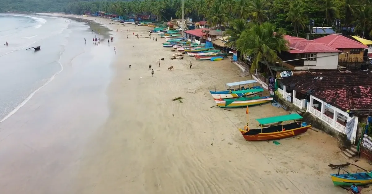 Panoramic view of Palolem Beach in Goa, featuring a wide sandy shore with colorful boats and beach shacks under an overcast sky.