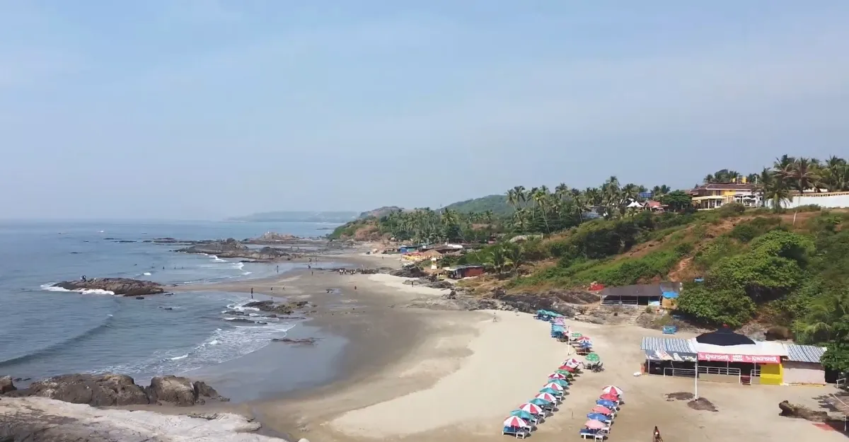 Varca Beach in Goa, showcasing its natural beauty and serene environment with a panoramic view of the coastline. The image captures the clear blue sky, golden sandy beach dotted with colorful umbrellas, and rocky terrain leading into the calm sea, exemplifying why Goa beaches are famous for their natural beauty and environment.