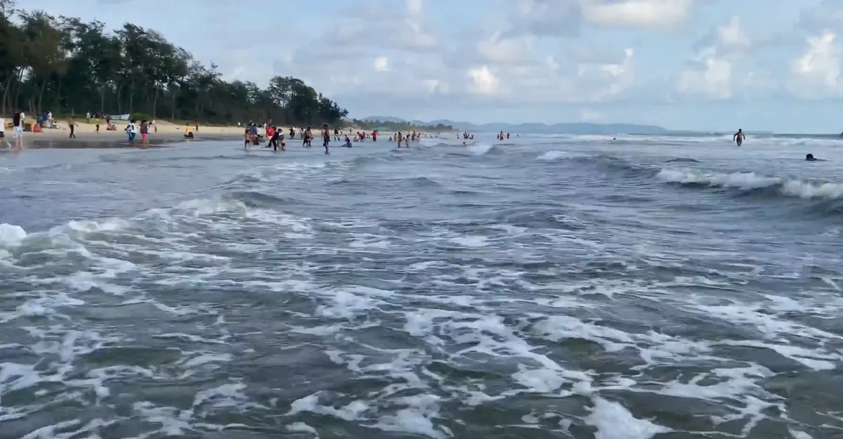 Scenic view of Varca Beach in Goa, India. The beach features a wide stretch of sandy shore with gentle waves lapping at the edge. Lush green trees line the back of the beach, and several visitors can be seen enjoying the serene atmosphere. The sky is partly cloudy, adding to the tranquil ambiance. In the distance, faint outlines of land or hills are visible on the horizon.