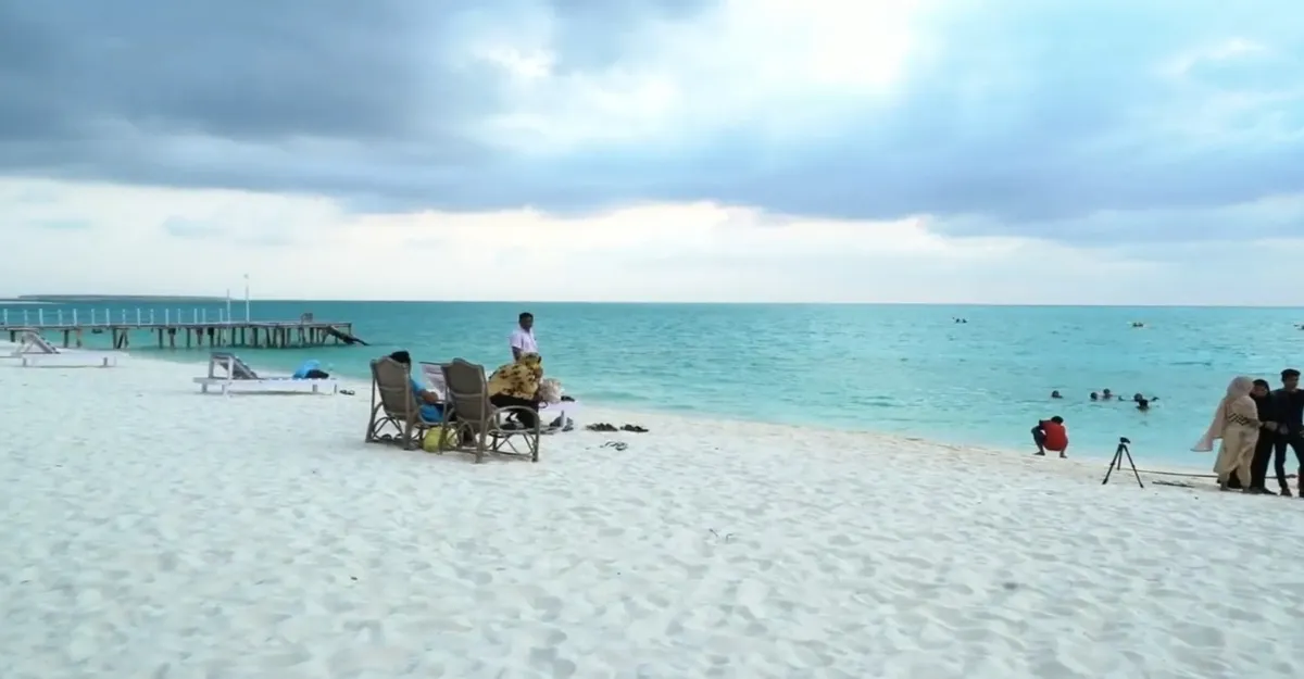 People and chairs on a white sandy beach with a wooden pier to the left. The ocean, reminiscent of Lakshadweep Beach, is a stunning turquoise under a cloudy sky.