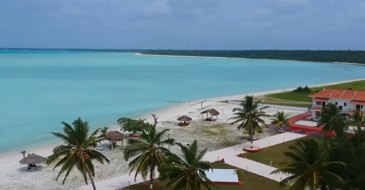 A scenic Lakshadweep beach offers turquoise waters and a sandy shore, with palm trees and thatched umbrellas dotting the landscape. A red-roofed building sits nearby, surrounded by lush green grass under a partly cloudy sky.