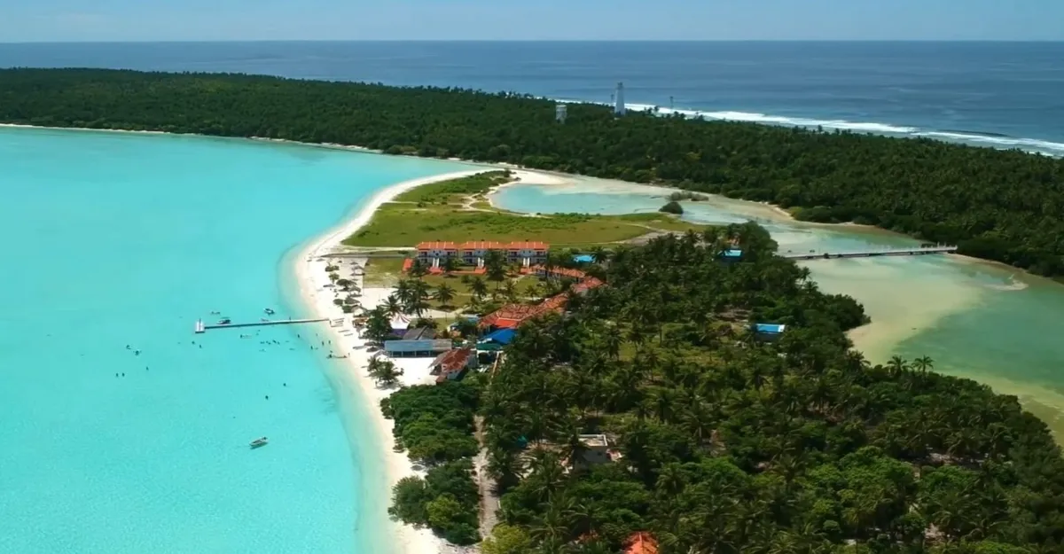 Aerial view of a tropical island in Lakshadweep with lush greenery, a white sandy beach, clear turquoise water, and small buildings near the shore.