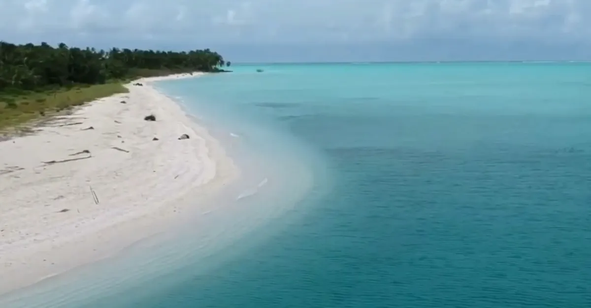 A serene Lakshadweep beach with white sand and clear turquoise water stretches along the coastline. The sky above is light blue with scattered clouds, while lush green vegetation lines the edge of this tranquil paradise in the distance.