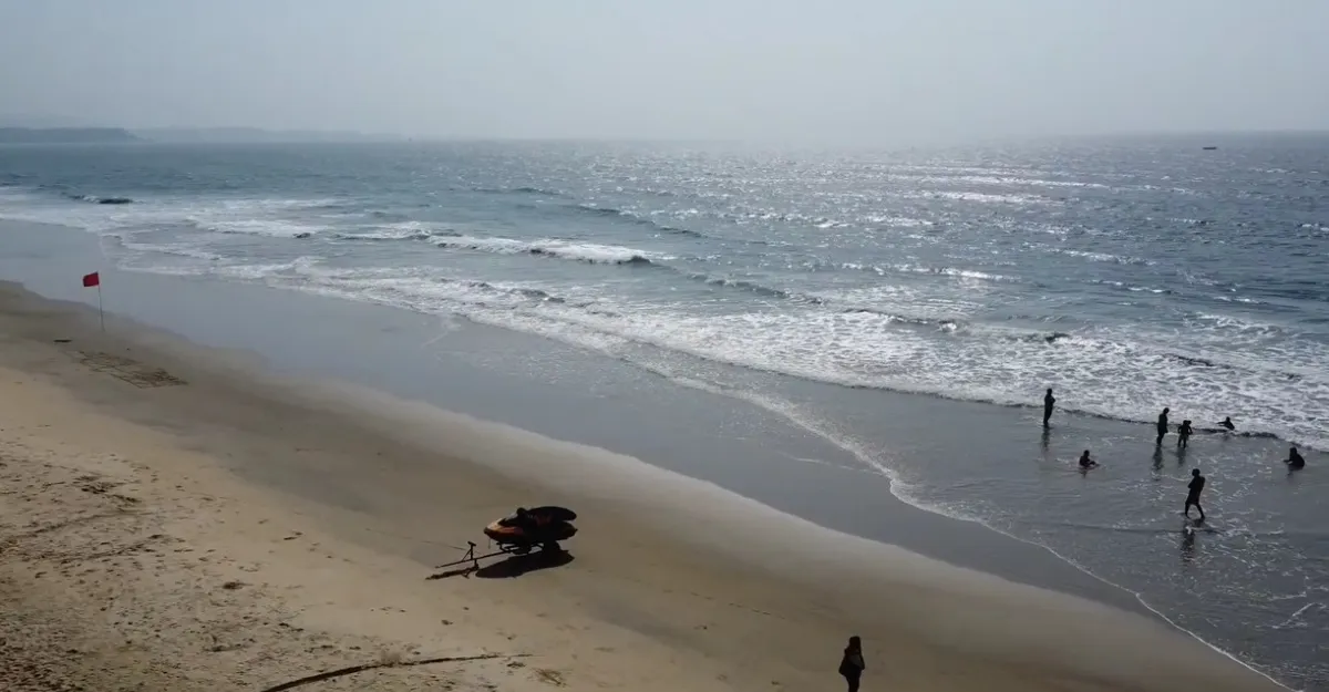 A serene view of Cavelossim Beach in Goa during the monsoon season, with gentle waves lapping the shore. A red flag is visible, indicating caution for swimmers, as a few people wade in the shallow water despite the seasonal risks. A lifeguard vehicle is stationed on the sandy beach, ready for emergencies. This scene highlights the question, "Is it safe to swim at Goa beaches during the monsoon season?" emphasizing the need for safety awareness due to strong currents during this time. The overcast sky and misty horizon add to the calm yet cautious atmosphere.