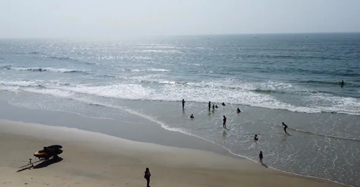 A serene view of Cavelossim Beach in Goa during the monsoon season, with gentle waves rolling onto the sandy shore. Several people are wading and swimming in the shallow water, enjoying the beach despite the monsoon's overcast sky. A jet ski is parked on the sand, indicating available water sports. This image raises the question, "Is it safe to swim at Goa beaches during the monsoon season?" as people appear to be taking precautions while enjoying the beach's natural beauty.