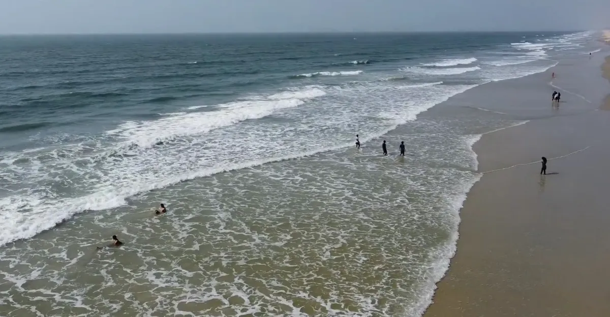 A view of Cavelossim Beach in Goa during the monsoon season, showing people swimming and wading in the shallow waters as waves gently crash onto the sandy shore. Despite the potentially strong currents typical of monsoon season, beachgoers are enjoying the ocean, prompting the question, "Is it safe to swim at Goa beaches during the monsoon season?" The image captures the lively atmosphere at the beach while highlighting the seasonal conditions that may impact safety.